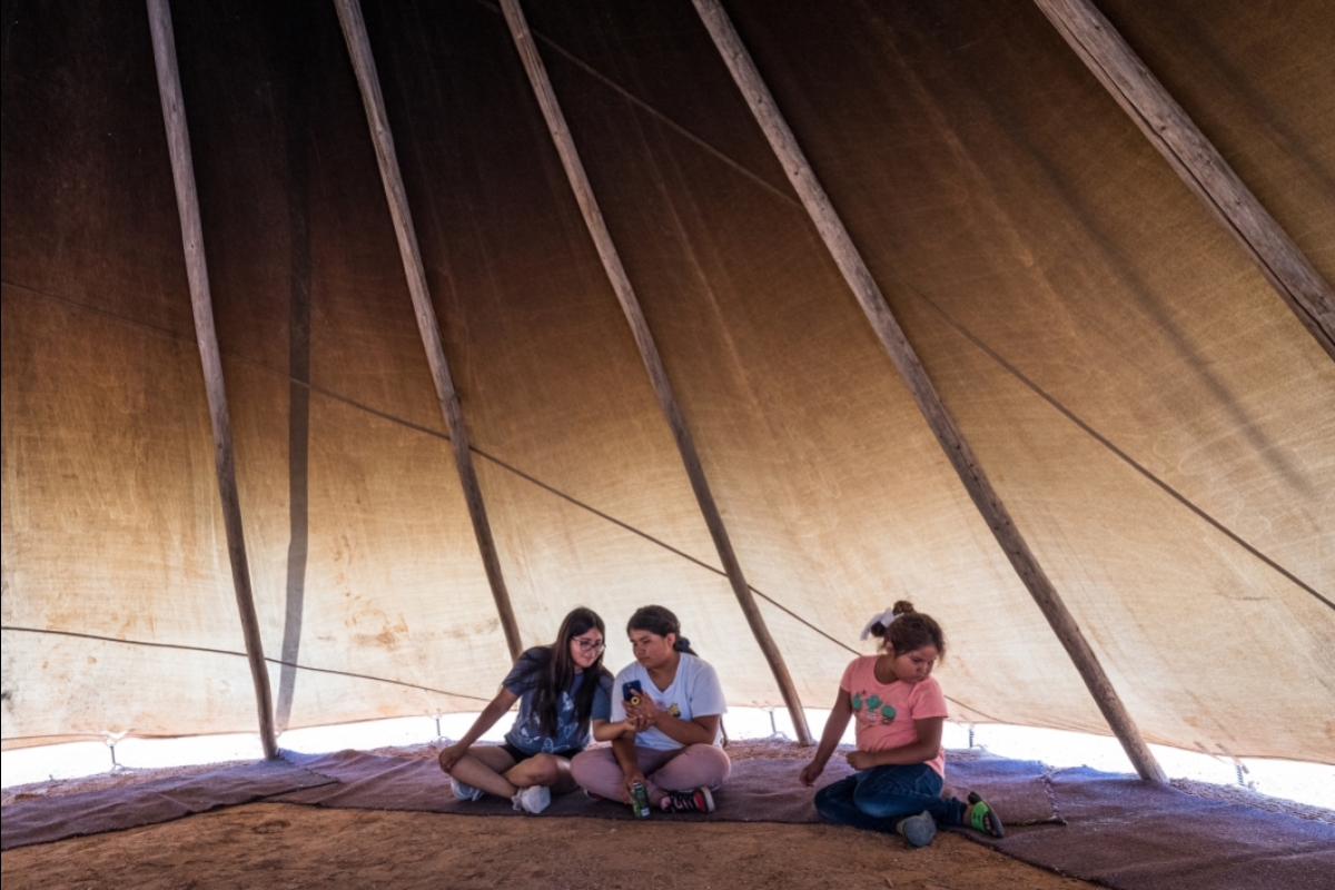 Three girls sit inside a teepee, the two oldest looking at a phone