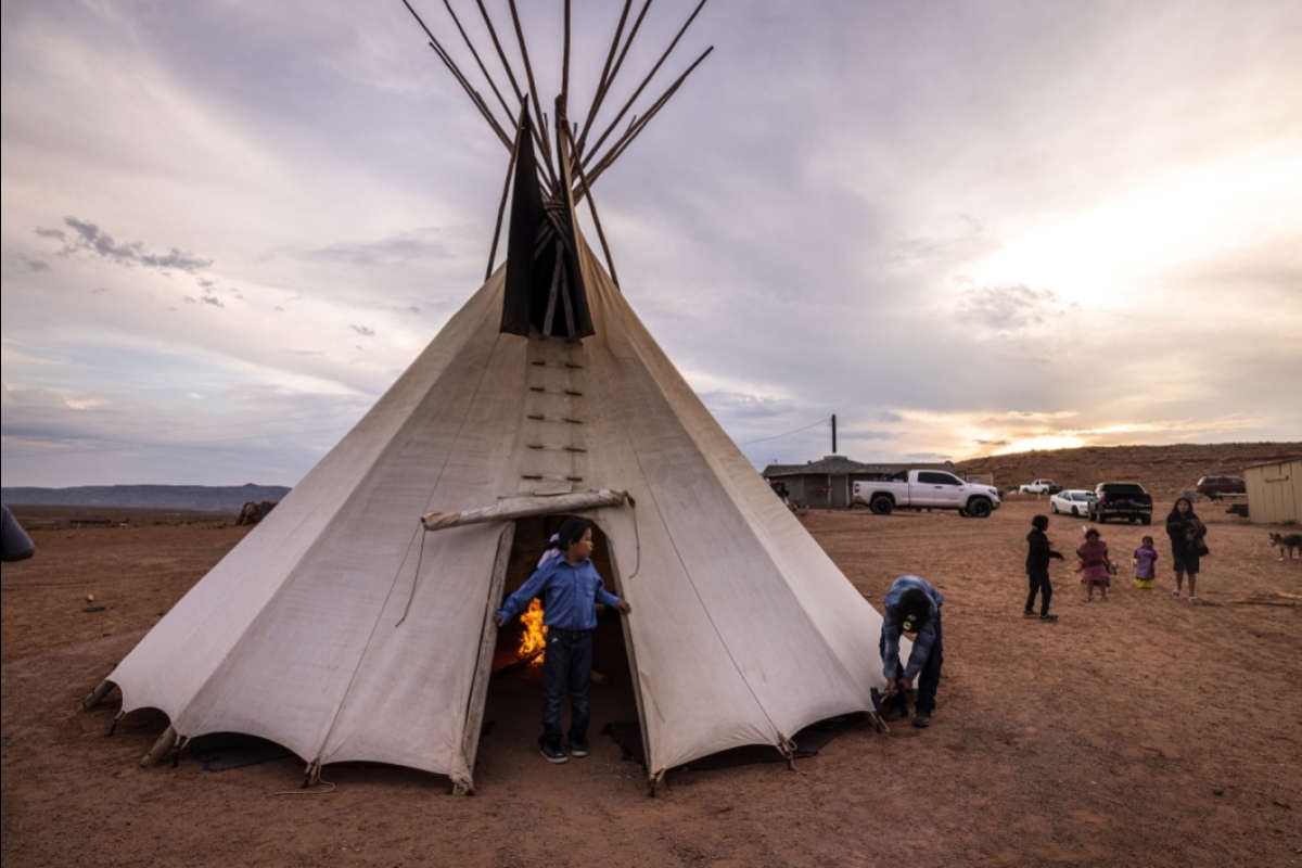 A man drives a stake at the base of a teepee while a boy stands in the doorway