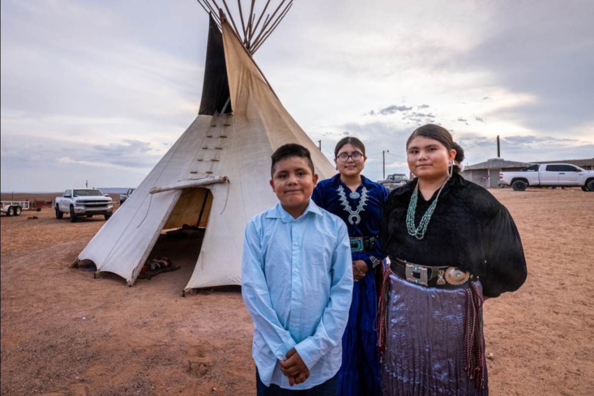 Three Navajo children in formal clothes stand outside a ceremonial teepee