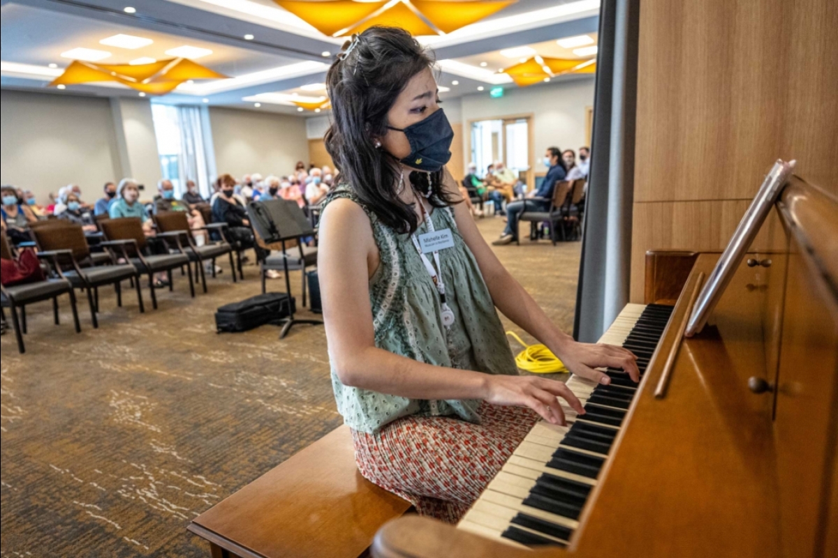 A woman plays the piano in front of a room full of people sitting in chairs
