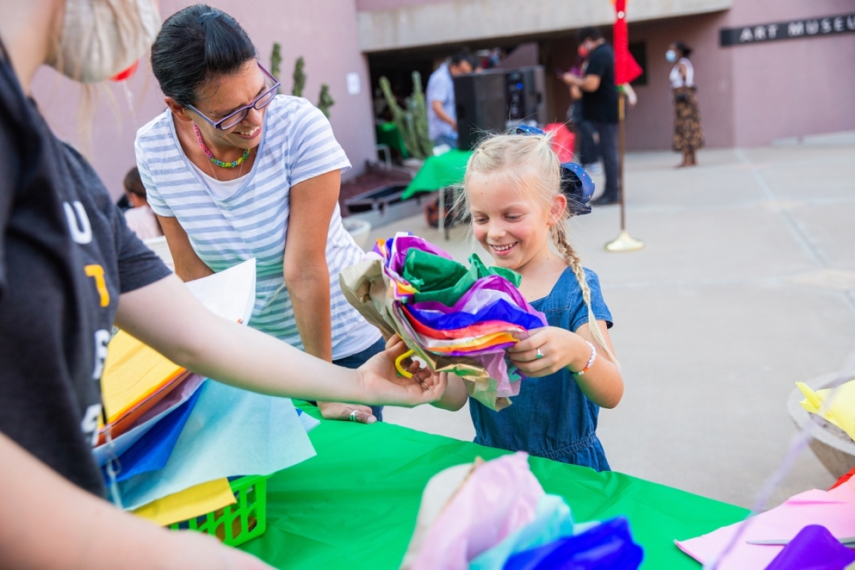 A little girl holds a stack of tissue paper at a craft table as her mom looks on and smiles
