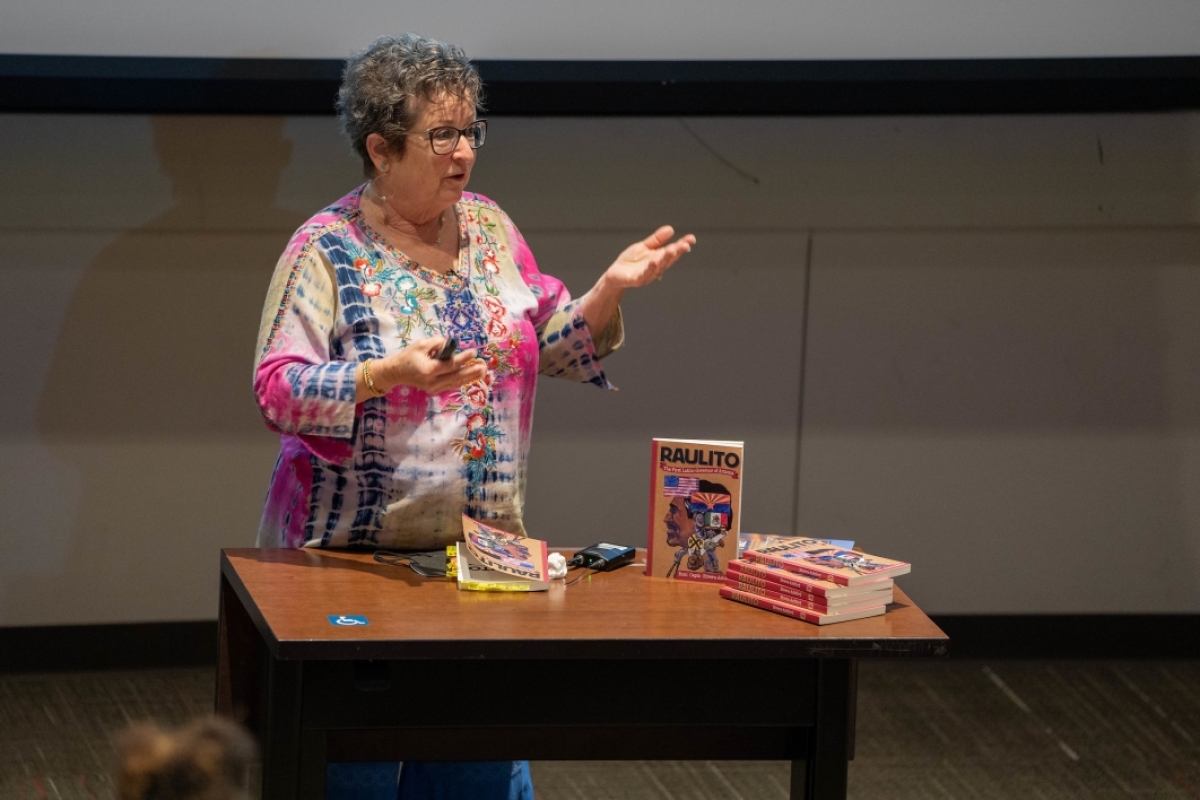 woman standing behind a table with books on it, gesturing with her arms as she speaks