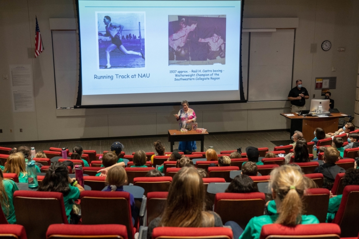 view from the back of an auditorium full of young students where a woman is speaking and pointing to a screen with photos on it