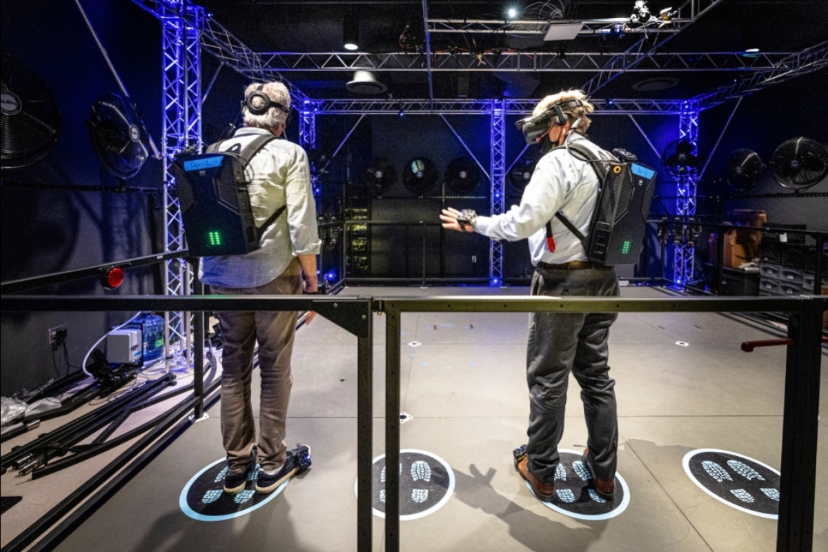 Two men seen from behind wear virtual-reality headsets, backpacks and hand-and-foot trackers.