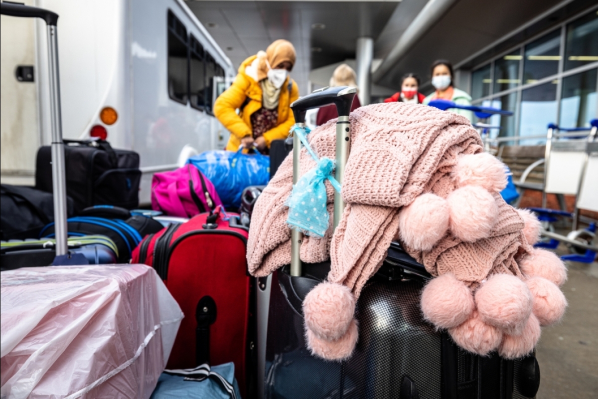 A pile of luggage sits on a sidewalk outside an airport, with women in headscarves standing behind