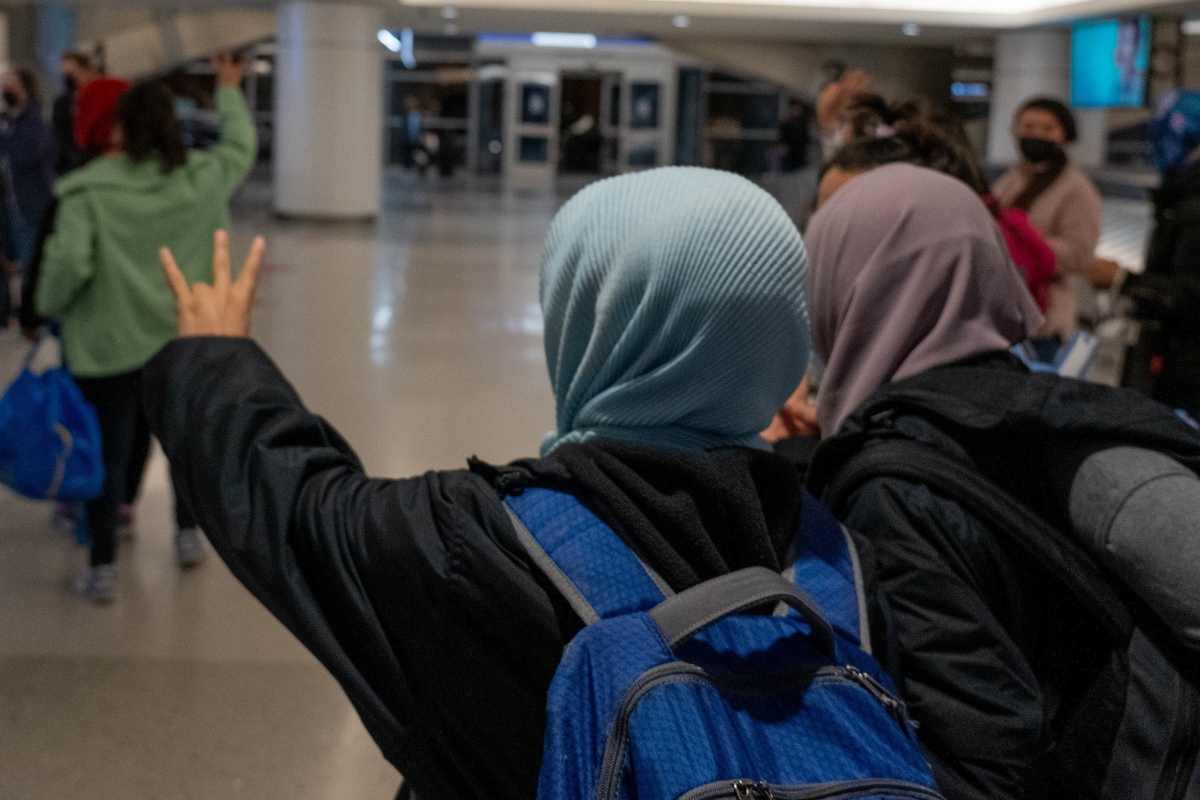 Two women in headscarves walk into an airport holding up the pitchfork gesture.