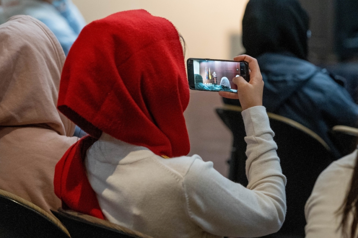 A woman in a headscarf films a speaker on her phone