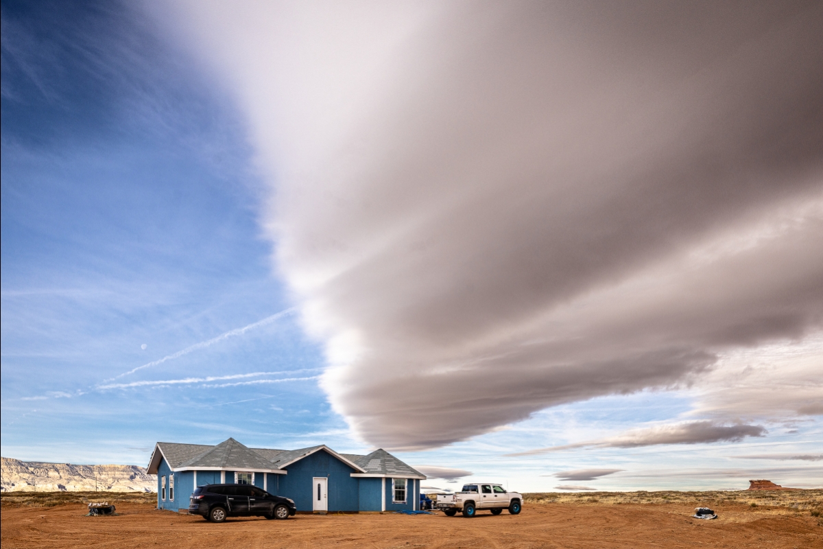 A blue house sits on a remote high desert landscape with no other buildings visible, and clouds stretching through the sky above