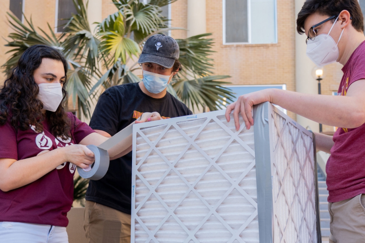 Three students wearing masks tape together air filters in the shape of a cube.