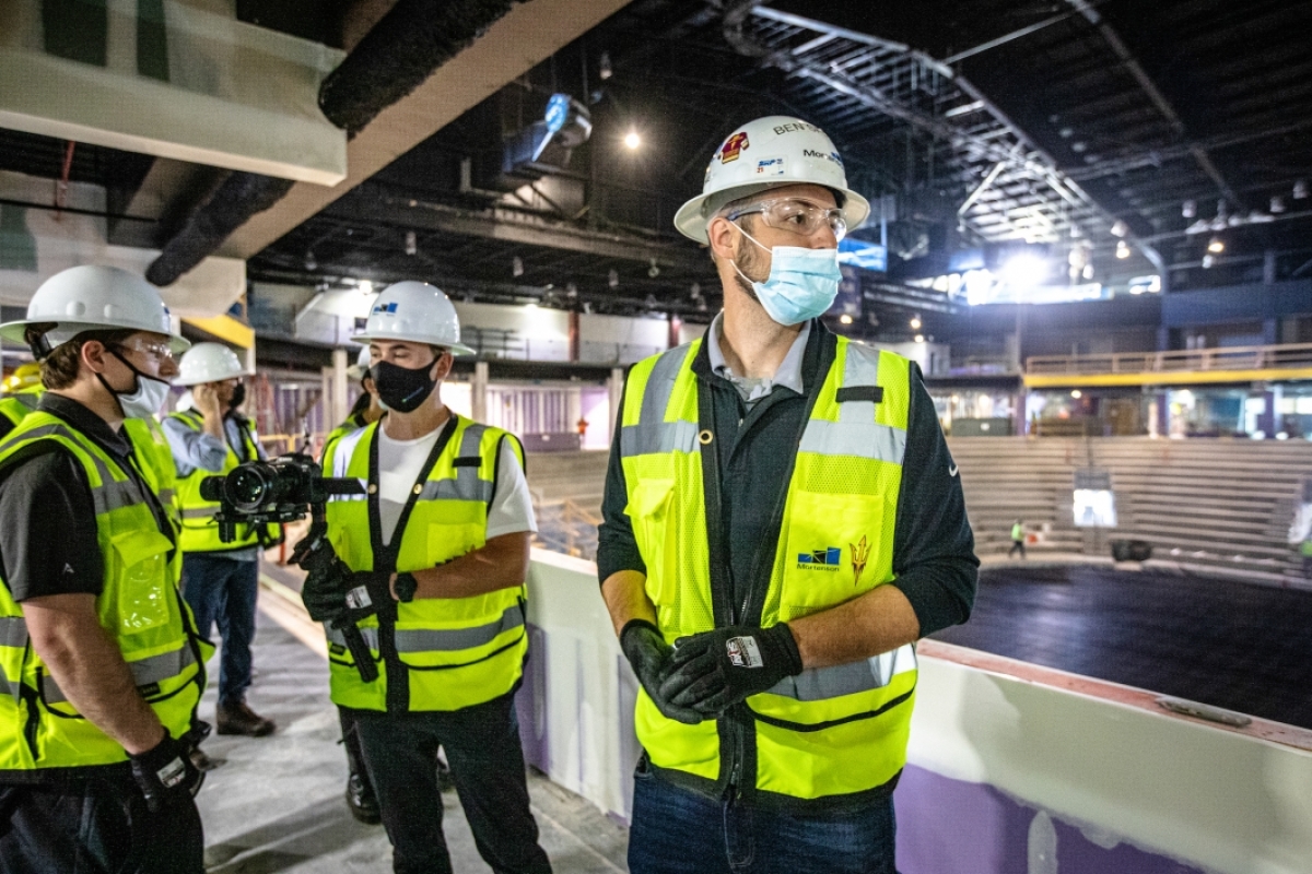 A man in a hard hat and neon yellow vest leads a media tour inside the new, under-construction ASU multipurpose arena