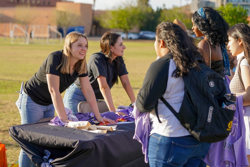Women standing behind a table answering questions from other women at an outdoor event.
