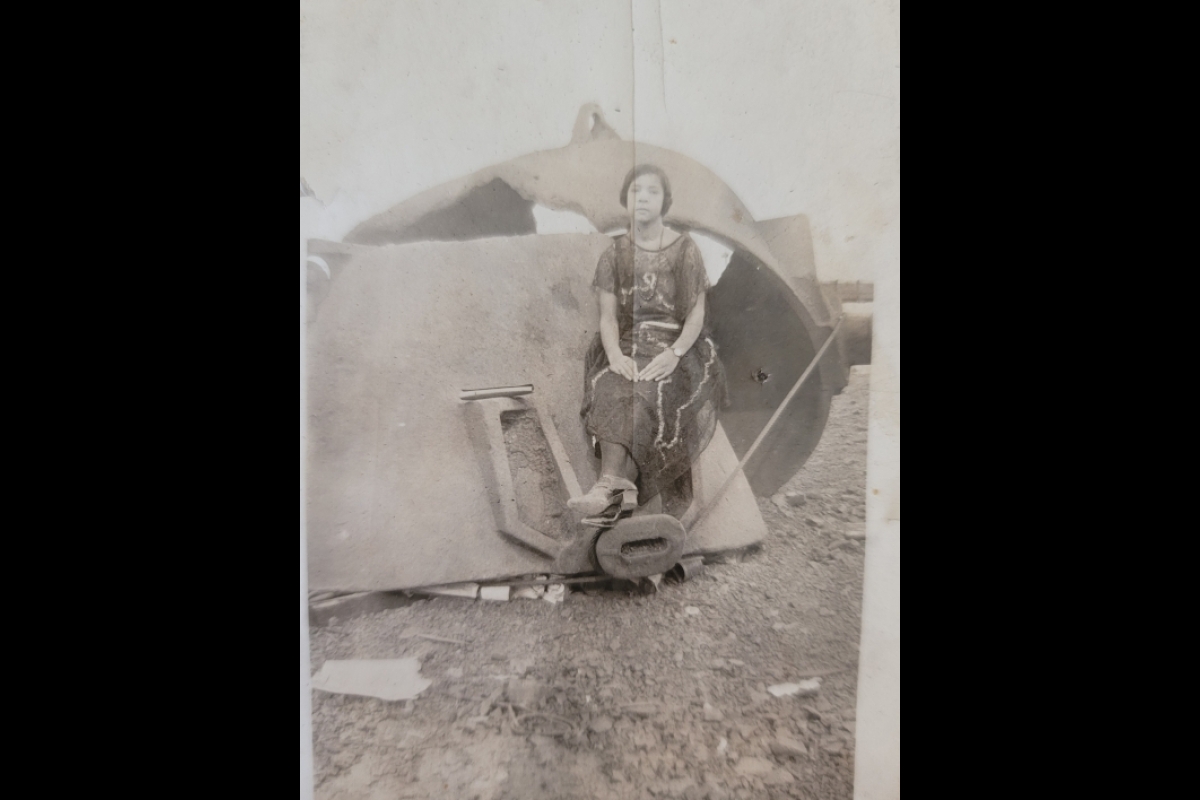 Old black-and-white photo of a young Black woman sitting on a peice of heavy machinery at a camp site.