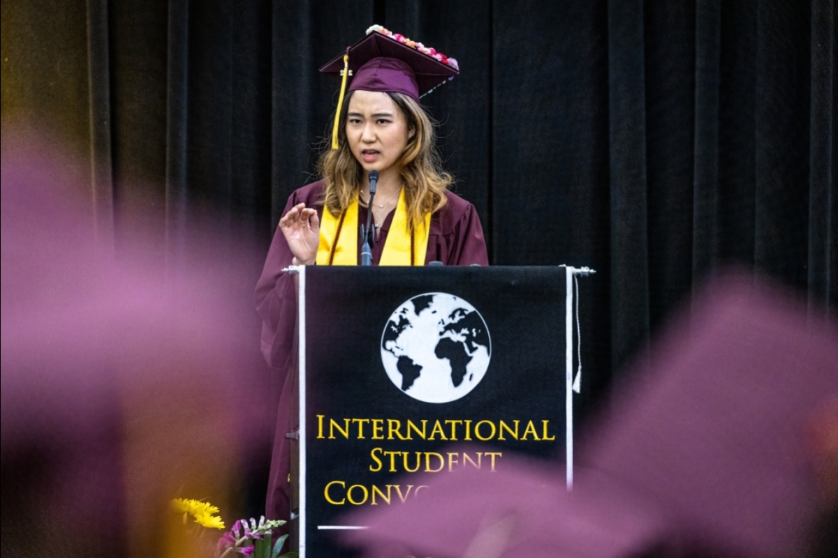 A woman in cap and gown speaks at a lectern with the words International Student Convocation on it
