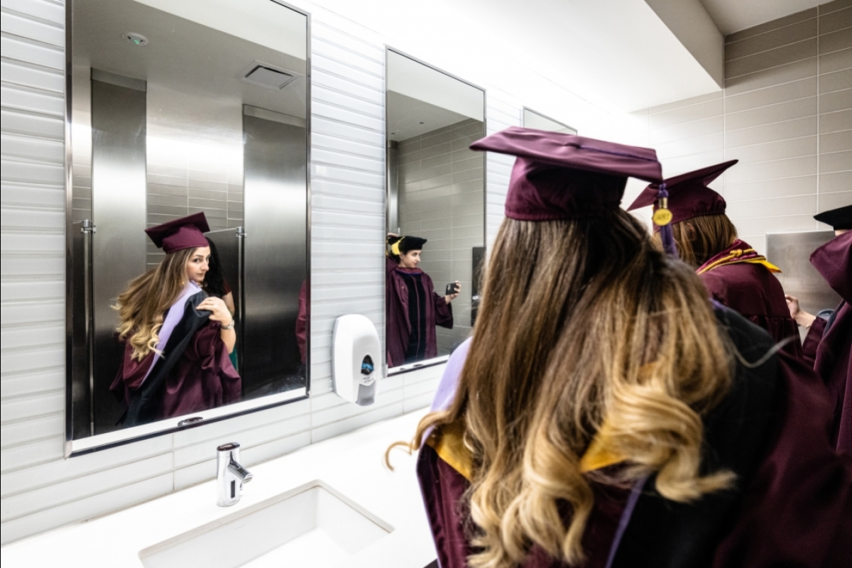 Women in graduation caps and gowns adjust their regalia looking into bathroom mirrors