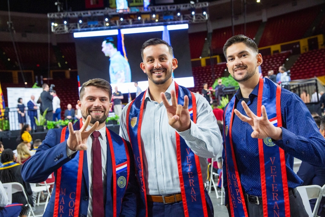 Three men wearing graduation ceremony regalia stand next to each other in an arena, smiling and making the ASU pitchfork symbol with their hands.