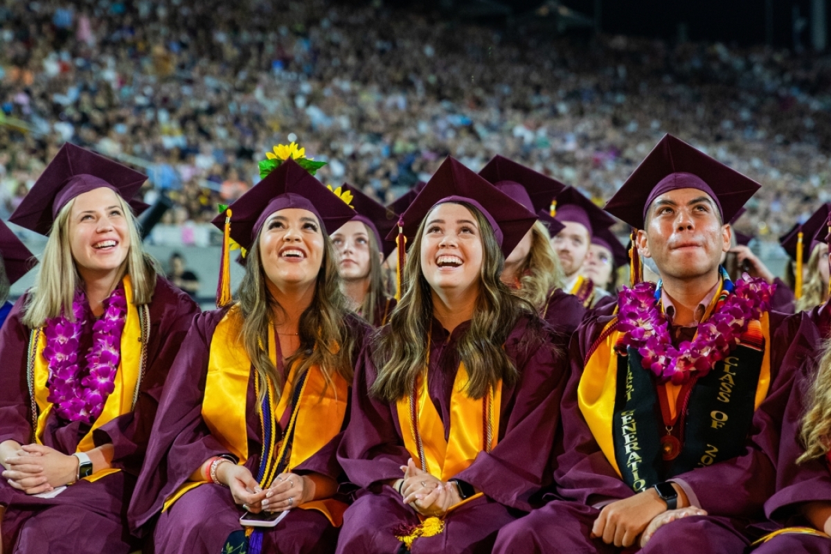 Three women and a man in graduation attire smile as they look up at a screen behind the photographer