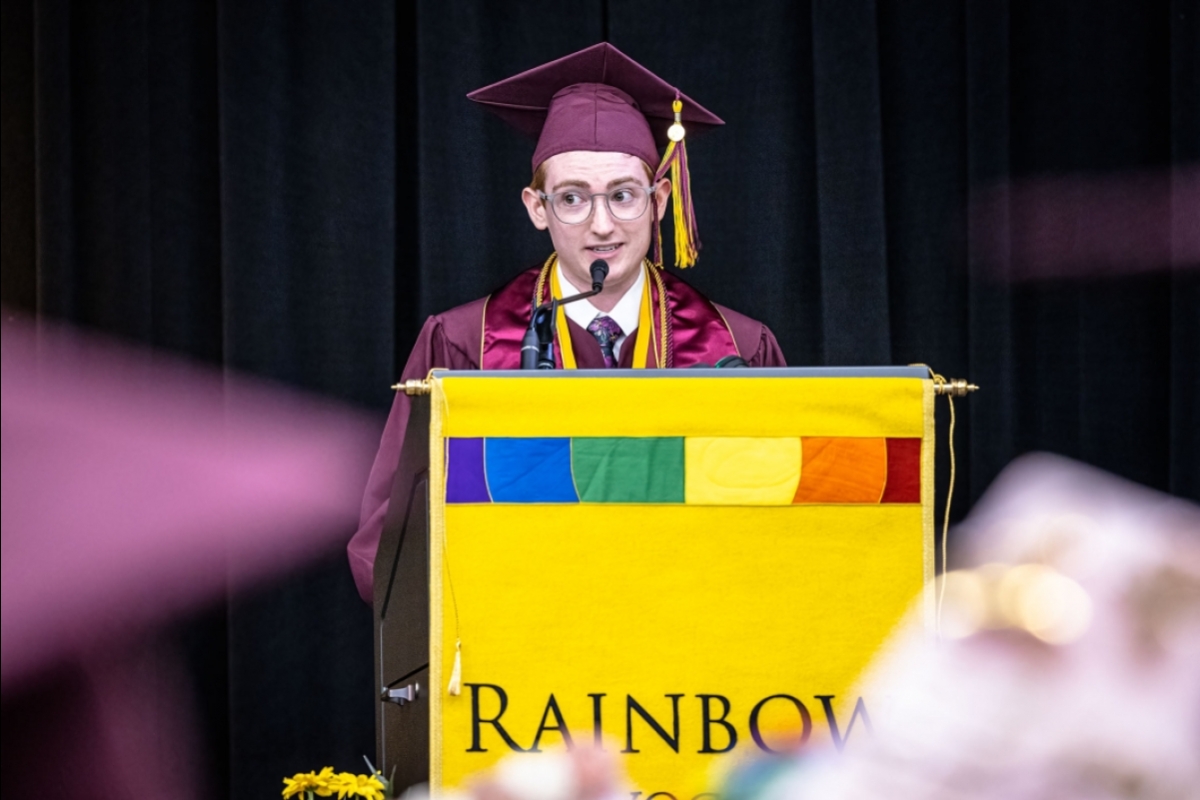 A young man speaks behind a lectern with a "Rainbow Convocation" sign on it.