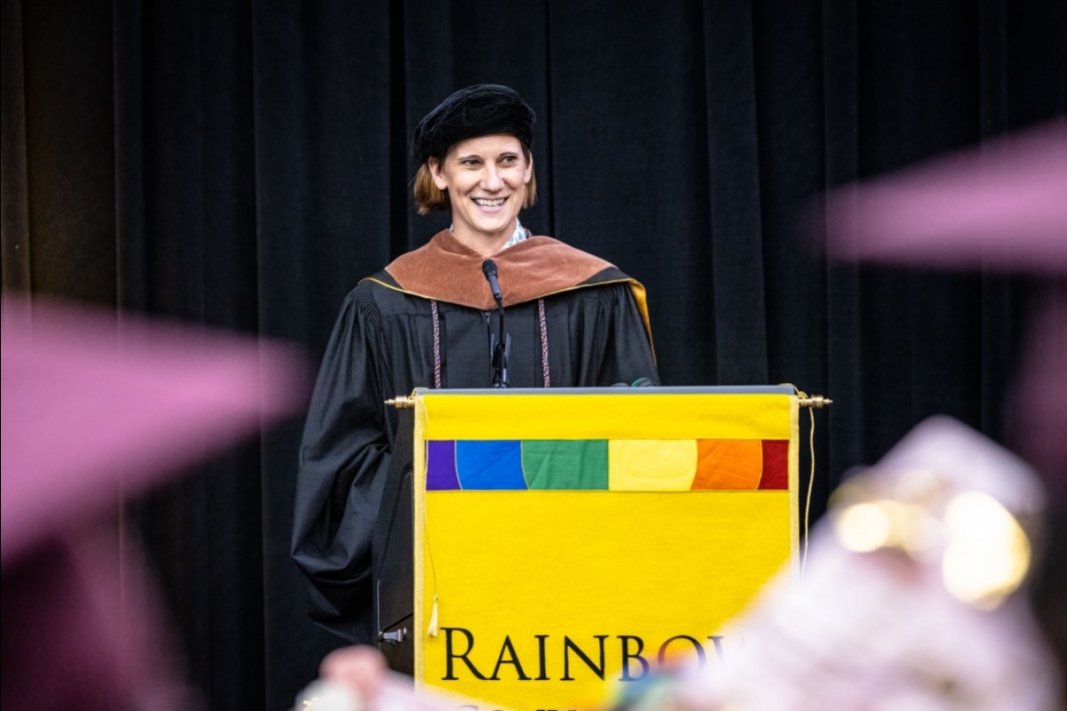 A woman speaks behind a lectern with rainbow colors and the word Rainbow Convocation on it