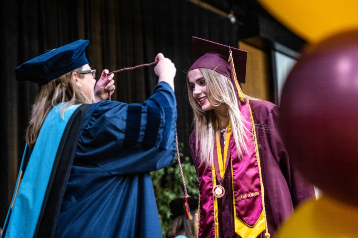 A faculty member in graduation regalia places a cord around a female student's neck