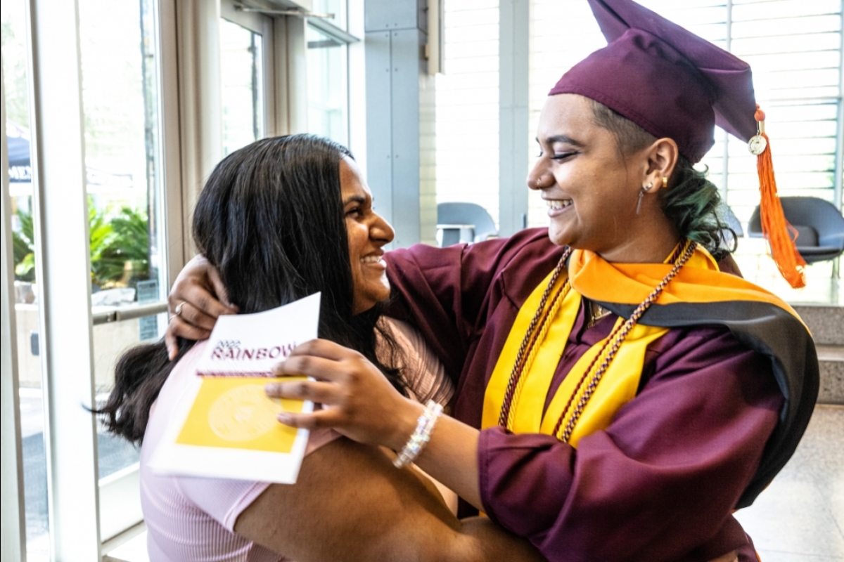 A student in graduation regalia hugs and looks at a friend with big smiles on both of them