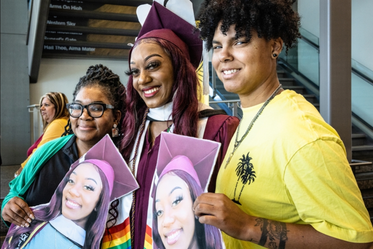 Two friends surround a woman in graduation cap and gown and pose for a photo, holding large printed signs of the graduate's face