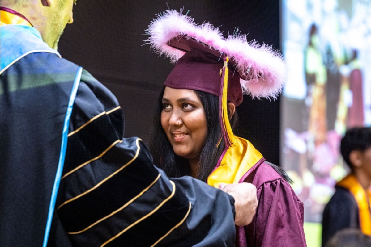 A woman whose graduation cap is ringed with a pink feather boa smiles as she receives a stole