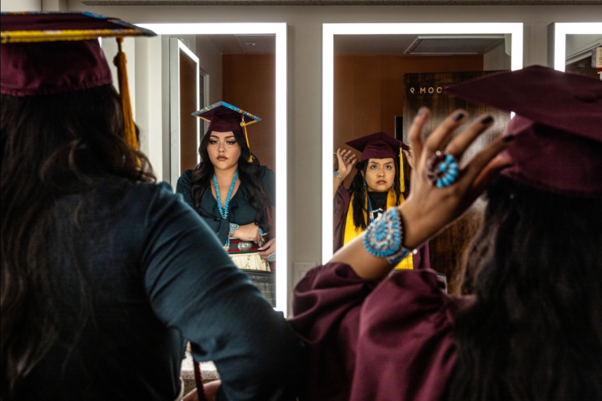Two women wearing caps and gowns and Native American jewelry look into dressing-room mirrors and make adjustments