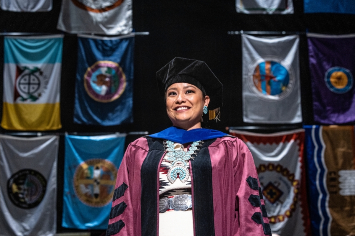 A woman in cap and gown smiles on a stage in front of a backdrop of tribal flags