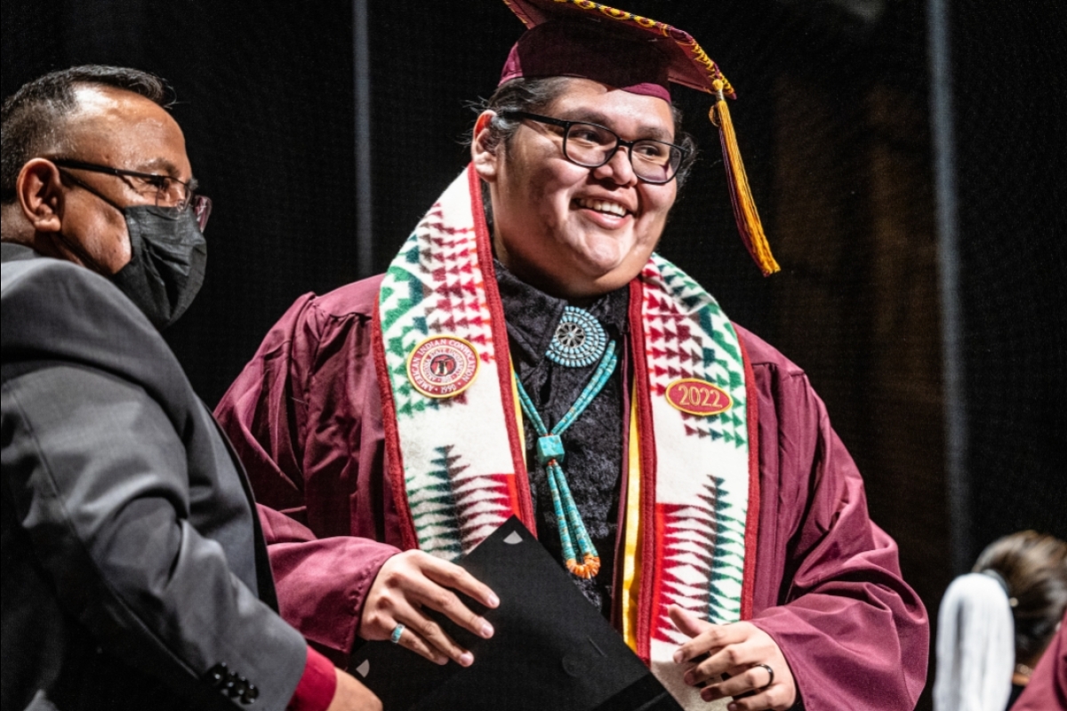 A tall young man in graduation cap and gown receives a stole with a Native American rug design
