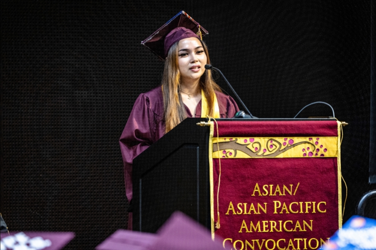 A young woman in graduation regalia speaks at a lectern