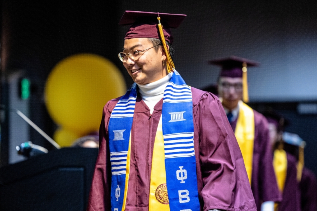 A young man in a graduation gown smiles as he crosses a stage