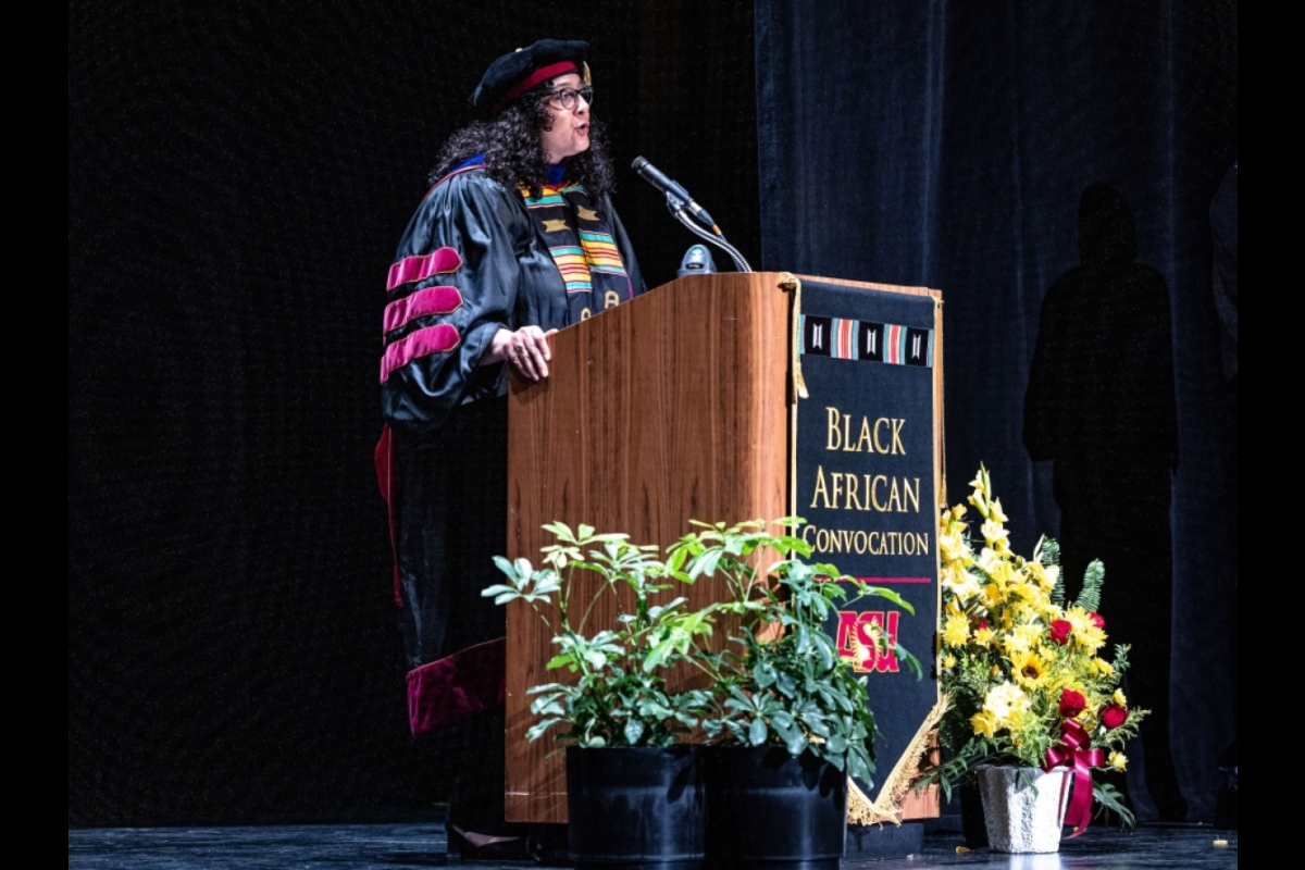 A woman in faculty academic regalia speaks at a lectern with a banner that reads Black African Convocation on the front