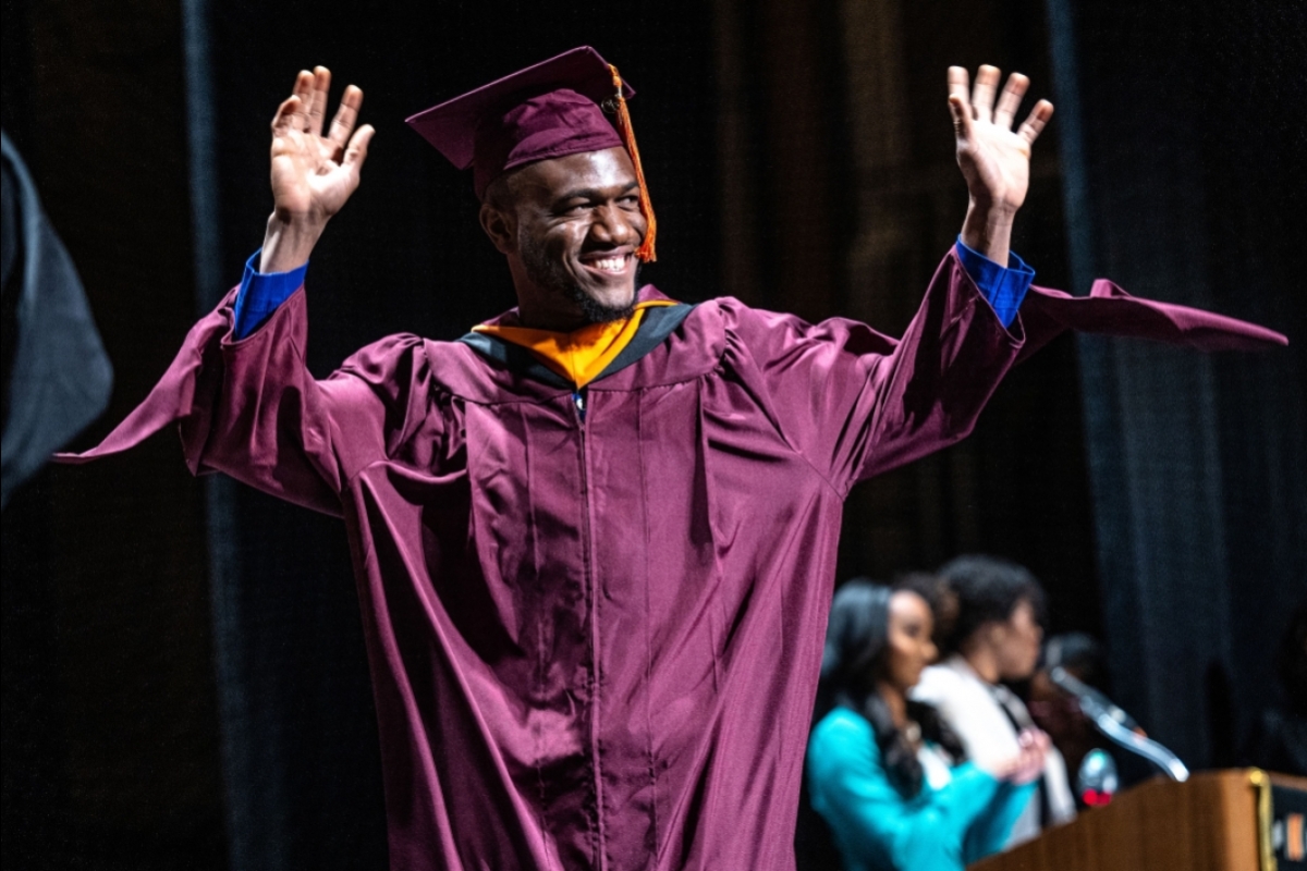 A man in graduation regalia waves from the stage
