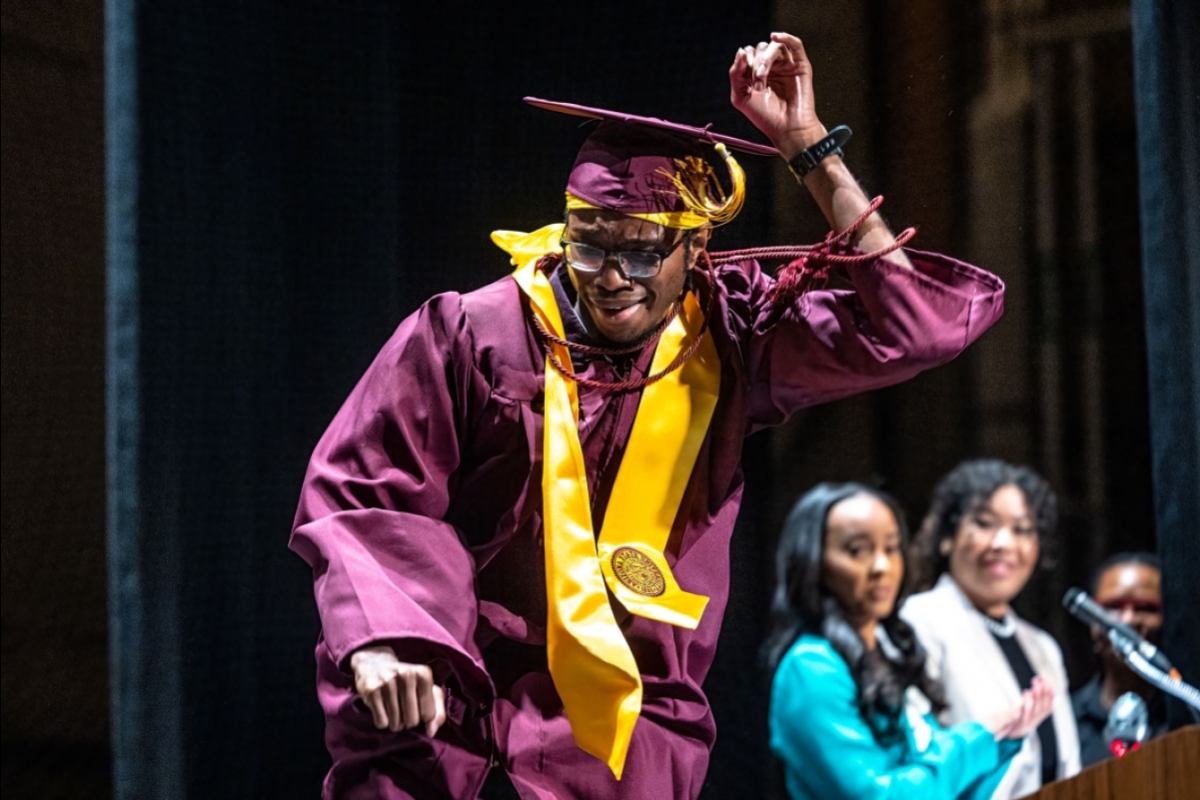 A man in graduation regalia dances across the stage to receive a stole