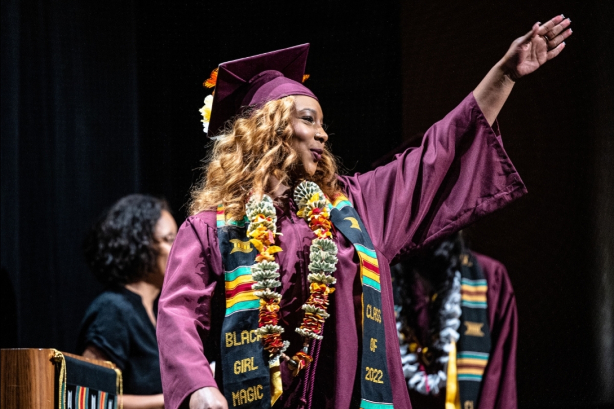 A woman in graduation gown and cap waves at the audience as she walks across a stage