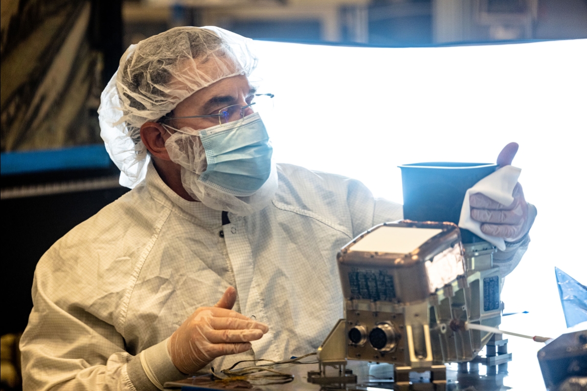A man in a lab coat, mask and hair net does a careful cleaning of a space imaging camera
