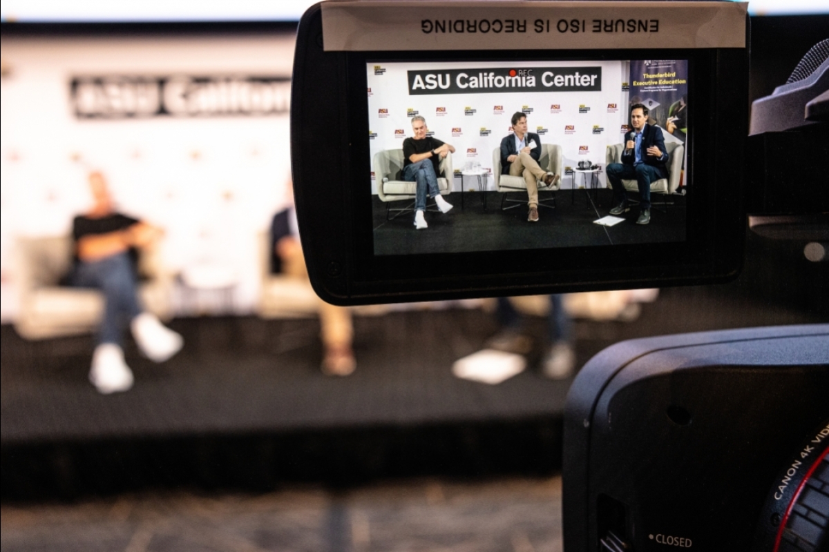 Three men on a stage are seen through the viewfinder screen of a video camera