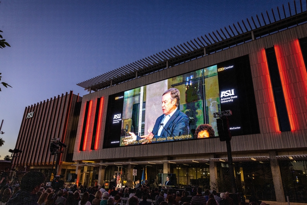 Exterior of a building with a large screen showing a man speaking to a crowd.