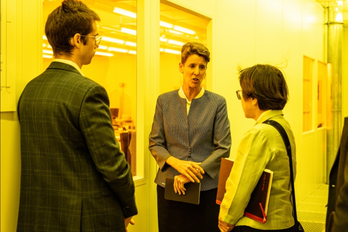 Two women and a man speak in a yellow-lit hallway outside a clean room lab