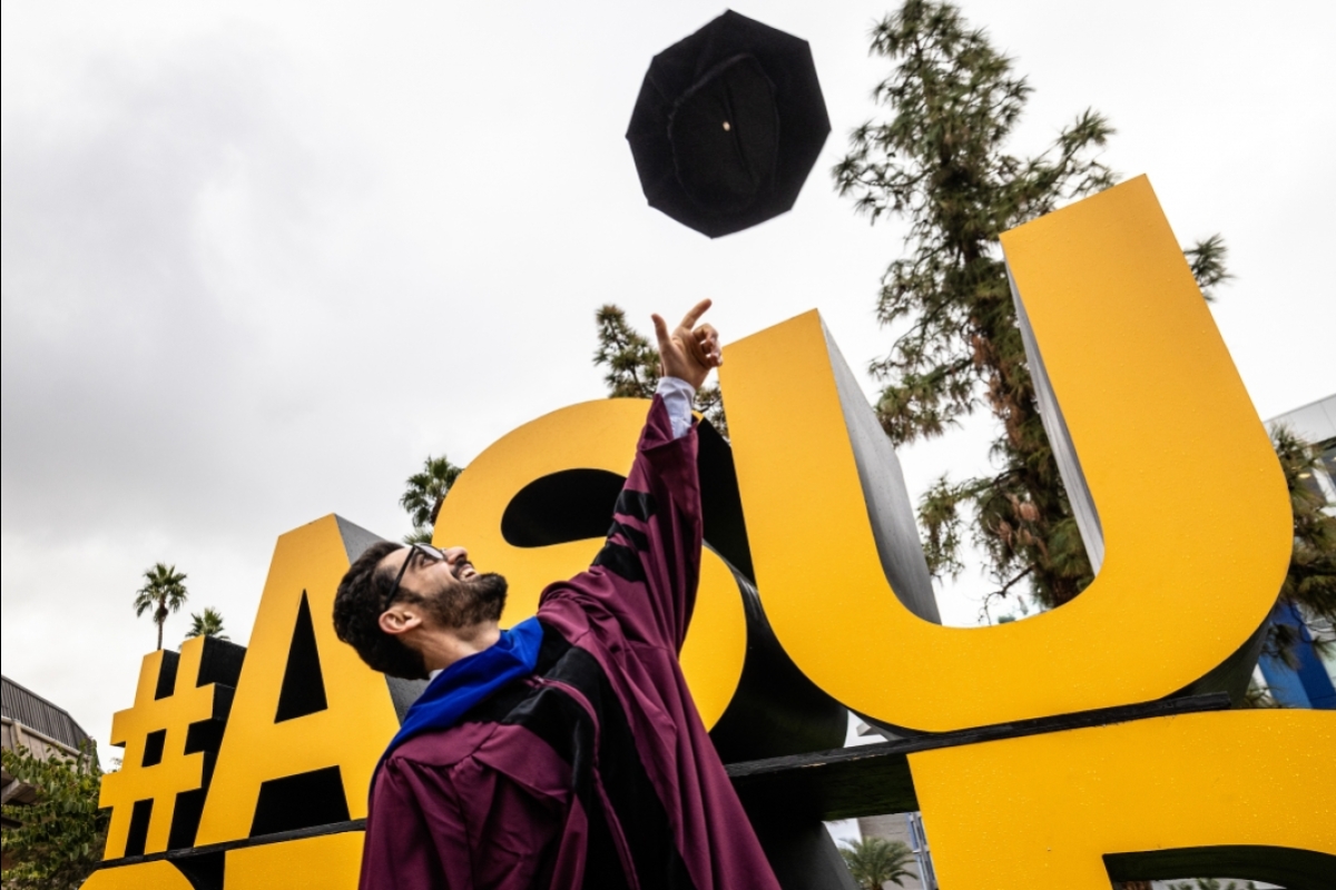 Grad tossing cap into air in front of large ASU graduation sign