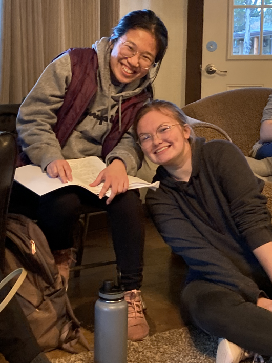 Two students smile for a photo while one holds a book open on her lap.