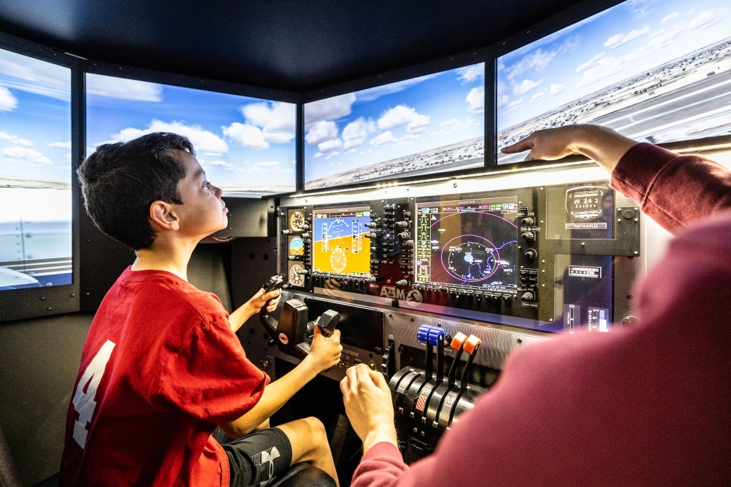 Young boy seated at an airplane flight simulator control panel gripping the yoke.