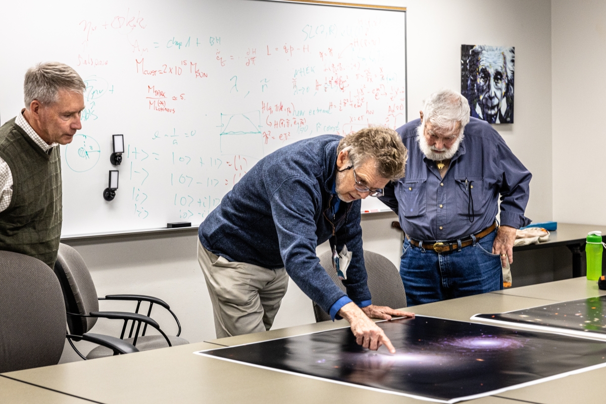 Three men leaning over a table looking at large images of galaxies.