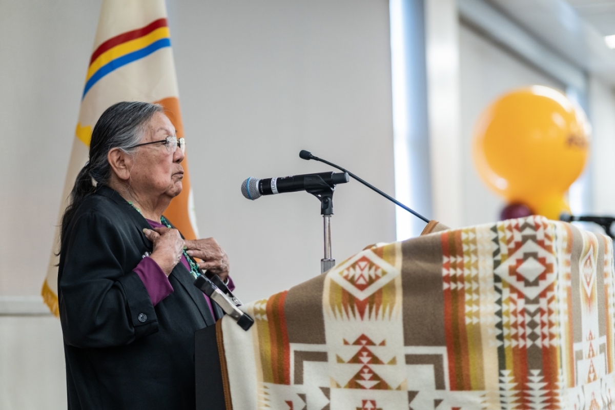 A woman speaks at a lectern covered in a Native American textile with a memorial photo on an easel to his right