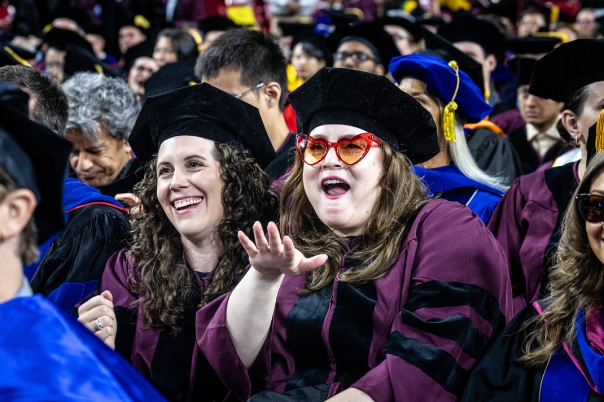 Two grads smiling, while one is wearing heart-shaped glasses, at commencement