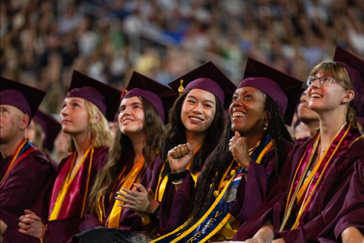 Group of sitting graduates in maroon caps and gowns watch a video play during commencement