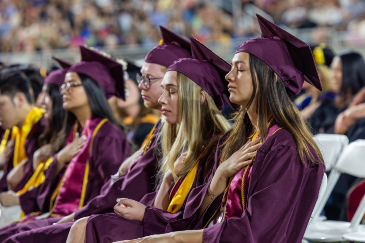 Grads in caps and gowns sitting during commencement closing eyes with hands on hearts