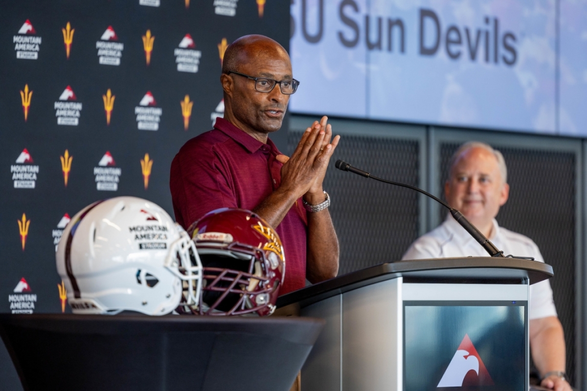 A man holds his palms together while speaking at a lectern next to football helmets