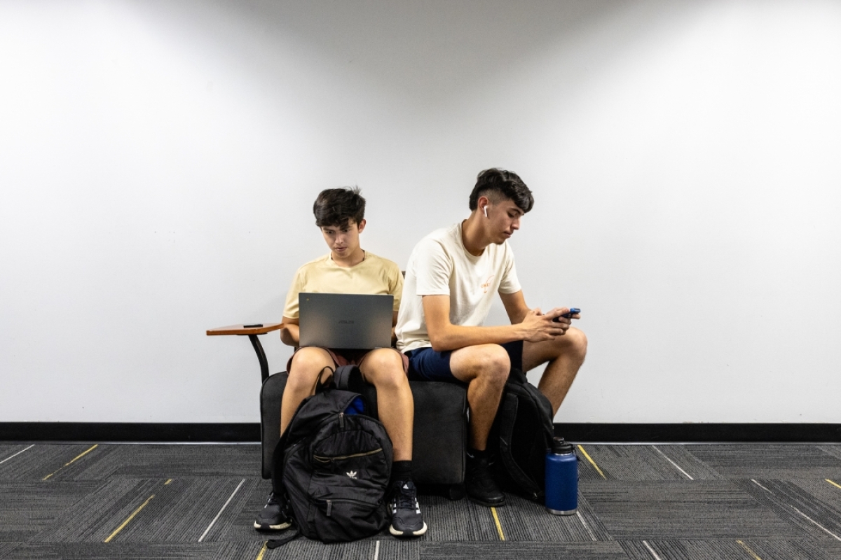 Two students sit back to back looking at computer and phone