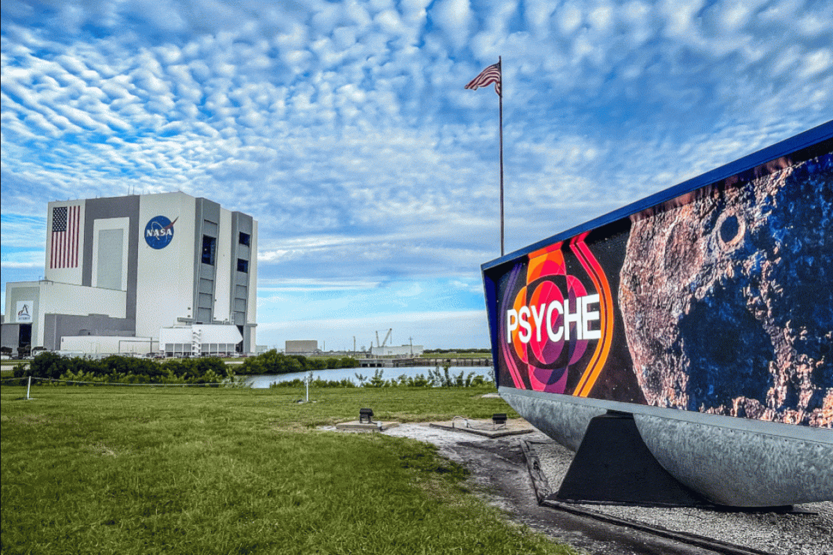 A sign with Psyche imagery in the foreground with the NASA Vehicle Assembly Building in the background